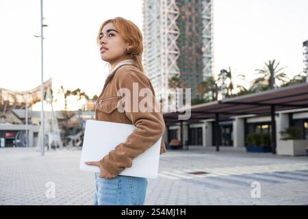 Eine gemischte Frau mit einem konzentrierten Ausdruck hält ein Notebook, während sie in einem städtischen Bereich steht und moderne Mode und Architektur präsentiert. Sie trägt ein Su Stockfoto