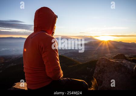 Rückansicht eines nicht erkennbaren Mannes, der auf einem Berg sitzt und den Sonnenaufgang beobachtet. Das Licht am frühen Morgen wirft warme Farbtöne über die Landschaft, die hell sind Stockfoto