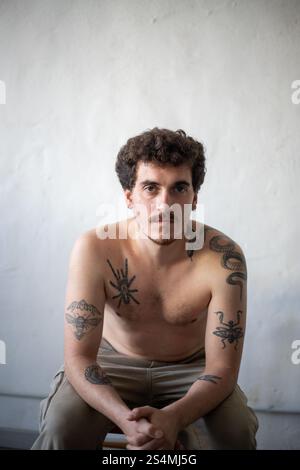 Ein junger Mann mit kurzem Bart und lockigem Haar sitzt entspannt vor einer weißen Wand und zeigt komplizierte Tattoos auf Armen und Brust Stockfoto