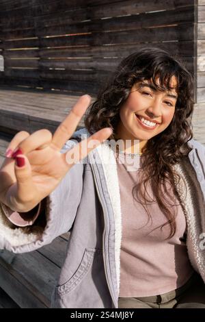 Junge Lateinfrau lächelt, während sie einen sonnigen Tag im Freien genießt, mit ihren Fingern ein Friedenszeichen blitzt und Freude und Positivität in einer lässigen Rela ausdrückt Stockfoto