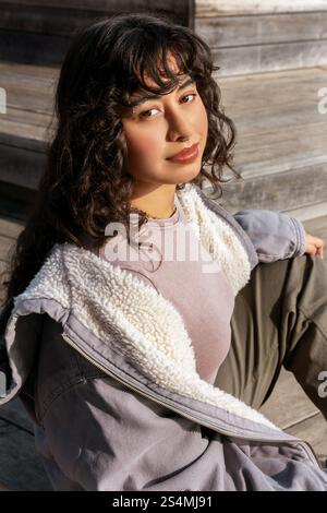 Eine junge Lateinfrau mit lockigen Haaren sitzt auf Holzstufen und genießt einen sonnigen Tag. Sie trägt eine gemütliche Jacke und legere Kleidung, die einen entspannten Stil ausstrahlt Stockfoto