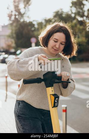 Eine junge Frau scannt mit ihrem Smartphone einen QR-Code auf einem Elektroroller und bereitet sich auf die Städtereise vor. Stockfoto