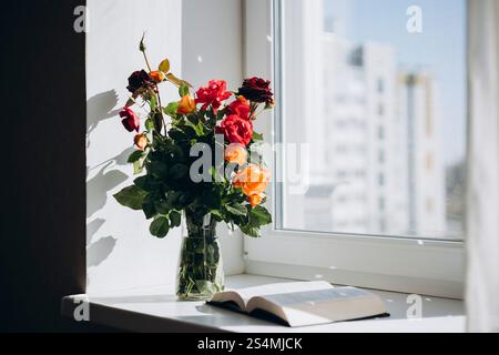 Ein lebhafter Rosenstrauß in einer Glasvase sitzt neben einer offenen Bibel auf einer sonnendurchfluteten Fensterbank und schafft eine ruhige und besinnliche Atmosphäre im Zimmer Stockfoto