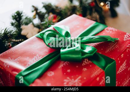 Ein wunderschön gewickeltes Weihnachtsgeschenk aus rotem Papier mit weißen Schneeflocken, gekrönt von einem luxuriösen grünen Band, festlichem Grün und Lichtern Stockfoto