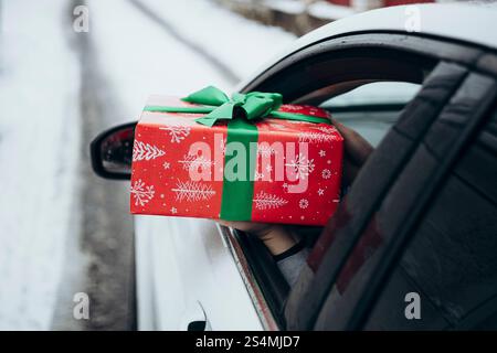 Eine Hand reicht ein wunderschön gewickeltes Geschenk mit rotem und grünem Urlaubspapier aus einem Autofenster. Das Auto parkt auf einer verschneiten Straße und sorgt für eine festliche Note Stockfoto