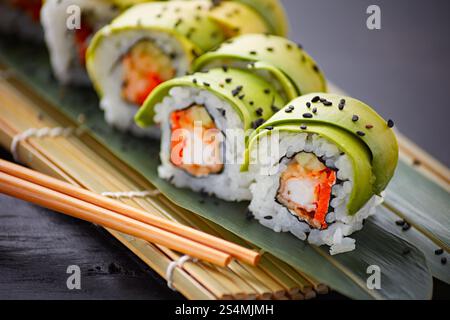 Köstliche Sushi-Brötchen, elegant in Avocadoscheiben gewickelt, serviert auf einer traditionellen Bambusmatte. Die Brötchen sind mit schwarzen Sesamsamen bestreut, A Stockfoto