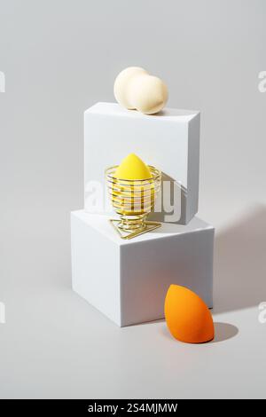 Drei Kosmetikschwämme in verschiedenen Farben ruhen auf minimalistischen weißen geometrischen Ständen vor einem weichen Hintergrund, die ihre lebendigen Töne und sm hervorheben Stockfoto