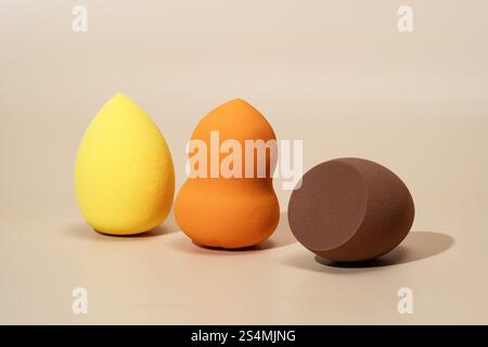 Drei farbenfrohe Kosmetikschwämme in Gelb, Orange und Braun sind auf einem neutralen Hintergrund angeordnet, ideal zum Auftragen und Verblenden von Fundamenten Stockfoto