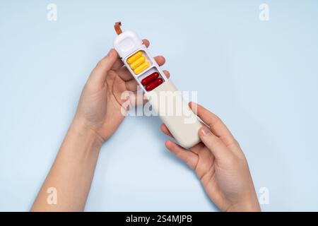 Von oben eine abgeschnittene, nicht erkennbare Hand, die eine weiße Pillbox mit verschiedenen farbigen Kapseln auf blauem Hintergrund hält. Stockfoto