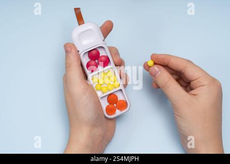 Zugeschnittene, nicht erkennbare Person, die eine gelbe Pille aus einem Handpillen-Organizer auswählt, gefüllt mit verschiedenen farbigen Tabletten, von oben gesehen gegen A Stockfoto