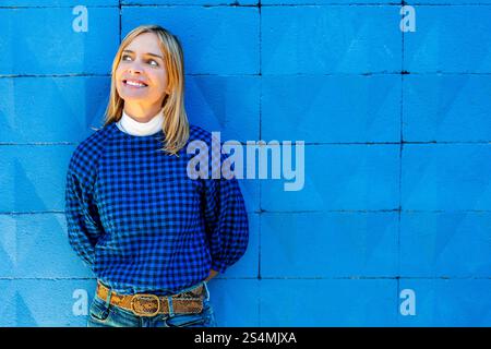 Eine fröhliche Frau lächelt in einem blauen karierten Hemd und lehnt sich an eine leuchtend blaue Wand. Sie schaut nach oben nach rechts, nicht d Stockfoto