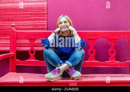 Eine fröhliche junge Frau in einem blauen karierten Hemd sitzt auf einer knallroten Bank, lächelt und blickt weg. Der Kontrast zwischen ihrer Freizeitkleidung und dem Stockfoto