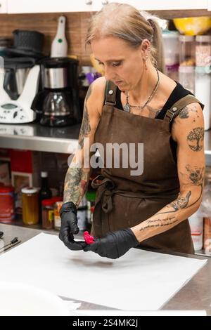 Ein erfahrener Handwerker in einer Bäckerei, trägt eine Schürze und Handschuhe, arbeitet akribisch an einem Stück Gebäck. Die Küche ist mit einer Vielzahl von Backwerkzeugen gefüllt Stockfoto