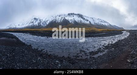 Ein dramatischer Blick auf einen schneebedeckten Berg, der sich über ein felsiges Flussbett im isländischen Hochland erhebt, mit dunklem vulkanischem Gelände und einer stimmungsvollen Überca Stockfoto