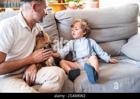 Ein fröhliches Vater-Sohn-Duo teilt einen warmen Moment mit ihrem kleinen Hund auf einem gemütlichen Sofa. Das Kleinkind in Hosenträgern streckt sich aus, um den verspielten Hund zu streicheln Stockfoto