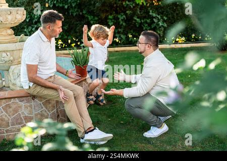 Fröhliche Szene eines schwulen Paares mit ihrem Sohn in einem üppigen Garten. Ein Vater ermuntert ihr springendes Kind, während der andere in der Nähe sitzt und alle smil teilen Stockfoto