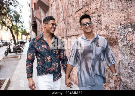 Ein fröhliches lateinisches Schwulenpaar läuft Hand in Hand durch eine lebhafte Straße in Madrid. Sie lächeln, tragen stylische Outfits und Sonnenbrillen und genießen die Sonne Stockfoto