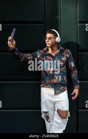 Ein stilvoller Mann mit Blumenhemd und Kopfhörern macht ein Selfie. Er trägt eine Sonnenbrille und zerrissene Jeans und steht selbstbewusst vor einem dunklen Hintergrund. Stockfoto