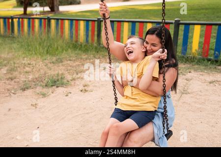 Eine liebevolle Mutter und ihr behinderter Sohn mit Zerebralparese teilen einen freudigen Moment auf einer Schaukel auf einem farbenfrohen Spielplatz. Die Szene fängt Wärme, Liebe ein Stockfoto