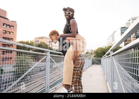 Ein glückliches, multiethnisches Paar aus LGBT genießt einen verspielten Moment auf einer Brücke in einer urbanen Umgebung, wobei ein Partner dem anderen einen Huckepack-Ritt gibt. Der Himmel i Stockfoto