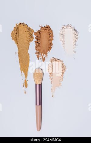 Draufsicht von drei verschiedenen Schattierungen von Gesichtspuder, die künstlerisch neben einem Make-up-Pinsel auf hellblauem Hintergrund verstreut sind. Stockfoto