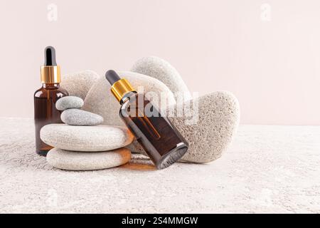 Zwei Serumflaschen aus braunem Glas mit goldenen Kappen sind kunstvoll zwischen glatten grauen Steinen ausgestellt, die ein ruhiges und Spa-ähnliches Ambiente zum Entspannen schaffen Stockfoto