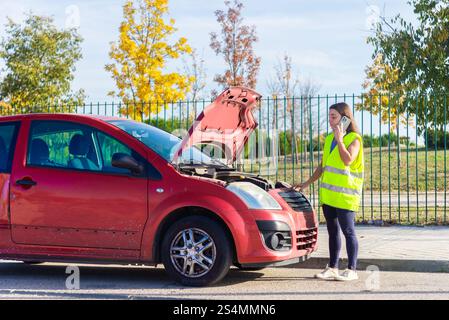 Eine Frau mit gelber Sicherheitsweste steht neben einem roten Auto mit geöffneter Motorhaube. Sie telefoniert am Telefon und bittet um Pannenhilfe auf einer sonnigen Straße Stockfoto