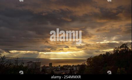 Ein atemberaubender Blick auf einen dramatischen Sonnenuntergang über dem Lake Leman, der goldene Strahlen durch stimmungsvolle Wolken über der Skyline von Lausanne wirft. Stockfoto