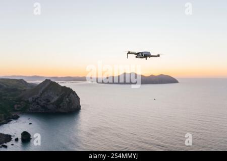 Eine Drohne fliegt anmutig über eine ruhige Küstenszene bei Sonnenuntergang. Das ruhige Meer und die Silhouetten entfernter Inseln bilden eine friedliche Kulisse von n Stockfoto