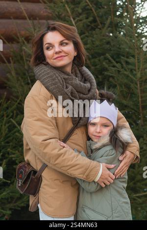 Eine Frau und ein junges Mädchen, die sich draußen in der Nähe eines Weihnachtsbaumes fest umarmen und in ihrer Winterkleidung Glück und Wärme ausstrahlen. Die Frau schaut weg Stockfoto