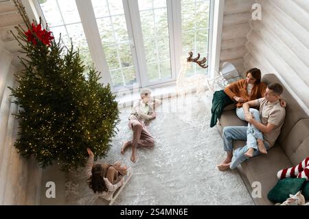 Eine Familie feiert Weihnachten in einem gemütlichen Zimmer mit einem großen, beleuchteten Weihnachtsbaum. Ein Paar sitzt auf einer Couch, zwei Kinder spielen in der Nähe; ein Mädchen reicht Stockfoto