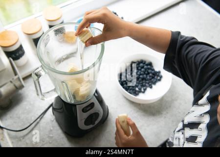 Ein Kind fügt Bananenscheiben in einen Mixer für einen frischen Smoothie hinzu. In der Nähe befindet sich eine Schüssel Heidelbeeren, die perfekt für ein nahrhaftes Frühstück oder einen Snack geeignet ist Stockfoto