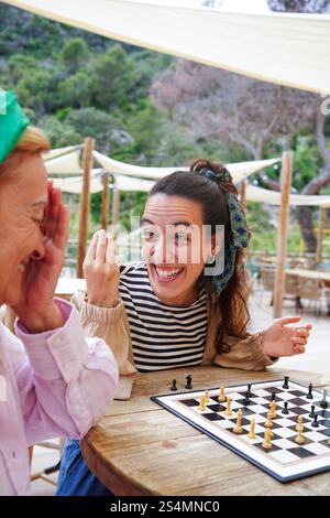 Mutter und Tochter teilen sich einen unbeschwerten Moment beim Schach außerhalb der Szene, fängt Lächeln und Strategie vor einem Hintergrund von Grün ein und fördern Stockfoto