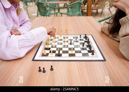 Mutter und Tochter spielen draußen auf einem Holztisch ein Schachspiel. Der Fokus liegt auf der strategischen Bewegung des Spielers in rosa grünen Stühlen und lässig Stockfoto