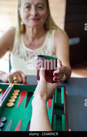 Nahaufnahme von zwei Personen, die Backgammon spielen, einer Hand einen Würfelbecher an die andere, während die Spielsteine auf dem Brett liegen und sich auf Interaktion und Freizeit konzentrieren Stockfoto