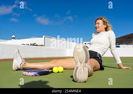 Eine junge Frau ruht auf einem Tennisplatz, sitzt mit ausgestreckten Beinen an einem sonnigen Tag. Mit geschlossenen Augen nach einem Spiel entspannend. Stockfoto