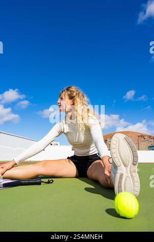 Frau mit blonden Haaren, in einer Stretching-Routine nach dem Spiel auf einem Tennisplatz. Sie sitzt bequem, streckt ihr Bein aus und lehnt sich sanft nach vorne, weg Stockfoto