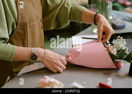 Mittlere Nahaufnahme von nicht erkennbaren weiblichen Händen, die pastellrosa Geschenkpapier mit kleinem Blumenstrauß anpassen Stockfoto