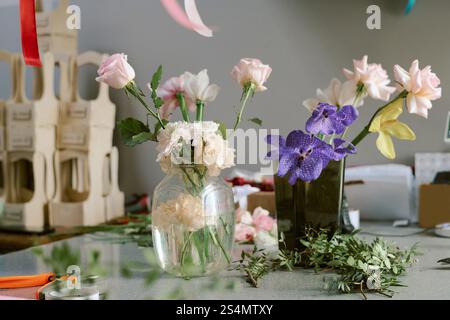 Mittlere Nahaufnahme von pastellrosa Rosen und violetten Orchideen in Krustenvase auf Tisch mit frischem und grünem Ruskus Stockfoto