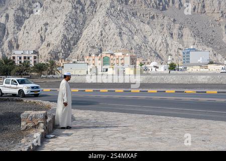 Khasab, Oman - 1. Januar 2025: Ein Mann in traditioneller omanischer Kleidung steht am Straßenrand vor dem Hintergrund rauer Berge und moderner Gebäude. Stockfoto