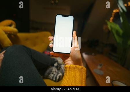 Smartphone Bildschirm Mockup Frau in gelben Pullover auf Couch halten Gerät zeigen Bildschirm warm Zuhause Innenleben Lifestyle grüne Pflanze Stockfoto