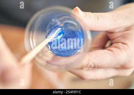 Nahaufnahme der Hände, die blaues Epoxidharz in einem klaren Kunststoffbecher mit Holzstab mischen. Stockfoto