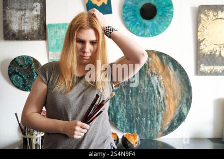 weiße Künstlerin mit blondem Haar, die Pinsel in einem Kunststudio hält, umgeben von abstrakten Gemälden. Stockfoto