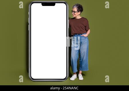 Ganzkörperfoto des hübschen älteren weiblichen Aussehens großes Smartphone tragen trendiges braunes Outfit isoliert auf khakifarbenem Hintergrund Stockfoto