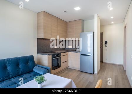 Moderne, minimalistische Küche mit Holzschränken, silbernem Kühlschrank und gemütlichem Wohnzimmer-Sofa in einem hellen Apartment. Minimalistische Küche und Wohnzimmer im Forts Stockfoto