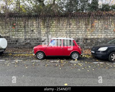Ein klassischer roter Mini Cooper parkt auf einer Straße zwischen zwei anderen Autos, mit einer Steinmauer im Hintergrund und gefallenen Blättern auf dem Boden. Stockfoto
