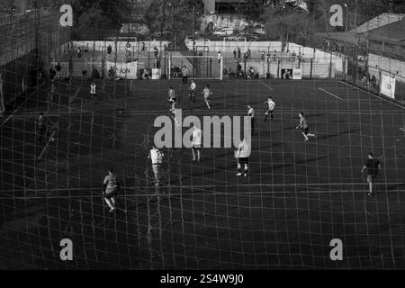 Schwarzweißfoto eines Fußballspiels im Freien auf einem städtischen Feld, durch Netznetz gesehen. Fängt Community, Teamwork und lokale Sportatmosphäre ein. Stockfoto