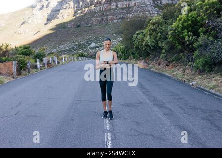 Die ganze Länge einer lächelnden Frau, die auf einer verlassenen Straße läuft und ihren Puls überprüft. Fröhliche Frau, die Smartwatches benutzt, während sie morgens spaziert. Stockfoto