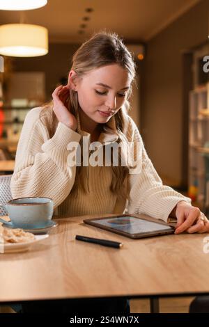 Blonde Frau, die ein digitales Tablet verwendet und im Café sitzt und online arbeitet, um ein neues Projekt am Computer zu erstellen Stockfoto