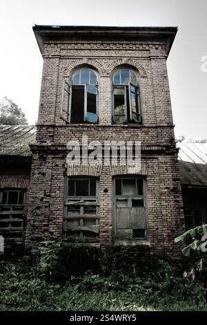 Altes, verfallenes Backsteingebäude mit zersplitterten Fenstern und dichter Vegetation, die die Umgebung erobert, was Mysterium und Verwüstung hervorruft. Stockfoto